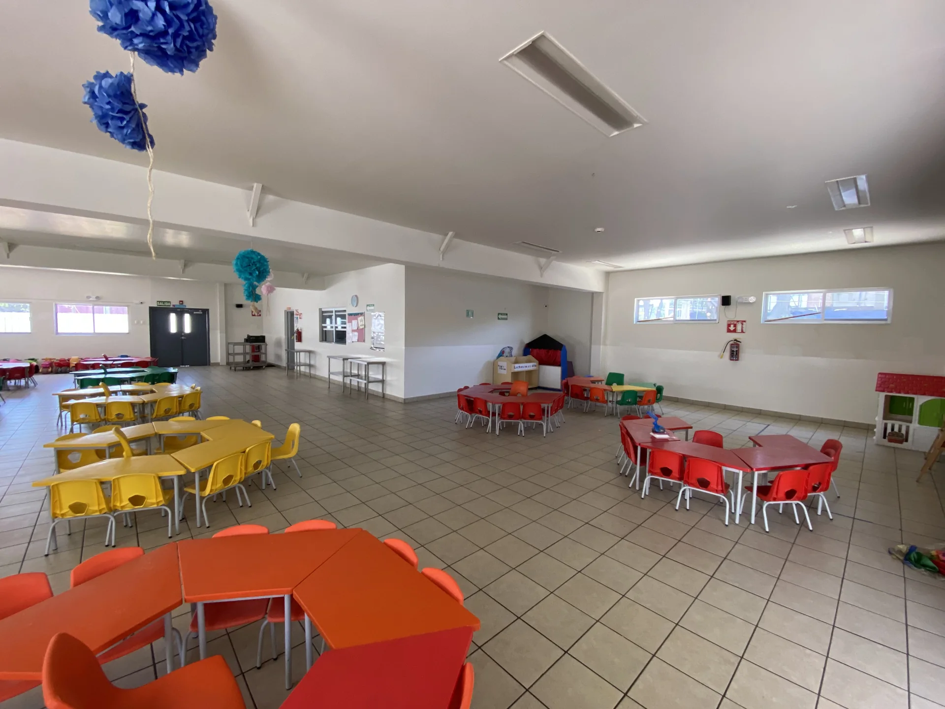 Sala de usos múltiples con mesas infantiles amarillas, rojas y verdes